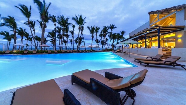 A swimming pool with lounge chairs, palm trees, and a building with a thatched roof. - Portblue Le Sivory Boutique Hotel (Punta Cana)