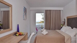 A hotel room with a bed, a desk with a fruit bowl, a chair, a mirror, and a view of the sea. - Bali Mare (Mylopotamos)