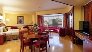 A hotel room with a dining area, a sofa set, a TV, and a large window with curtains.
