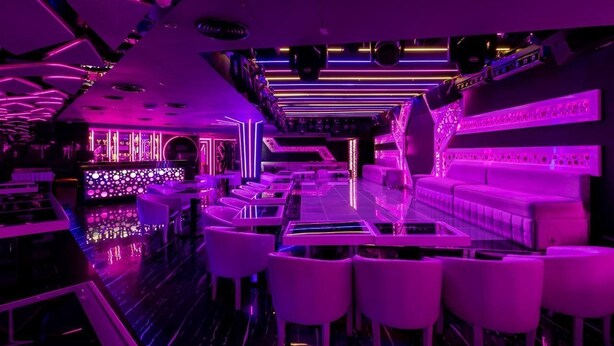 A modern bar with neon lighting, white bar stools, and a reflective floor.