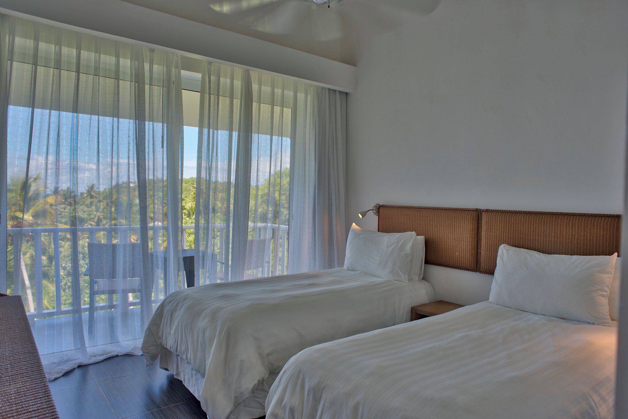 Une chambre d’hôtel avec deux lits, un balcon avec des rideaux légers et une vue sur la verdure.