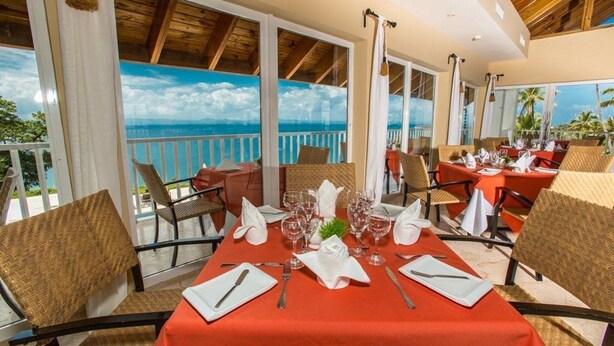A dining area with tables set for two, large windows with a view of the ocean, and a wooden ceiling. - Xeliter Vista Mare (Samaná)