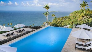 A swimming pool with lounge chairs and umbrellas, surrounded by palm trees and a view of the ocean. - Xeliter Vista Mare (Samaná)