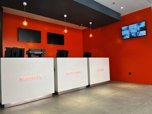 A reception area with a red wall, three white reception desks, black monitors, and a large TV screen. - Madrid Centro Atocha (Madrid)