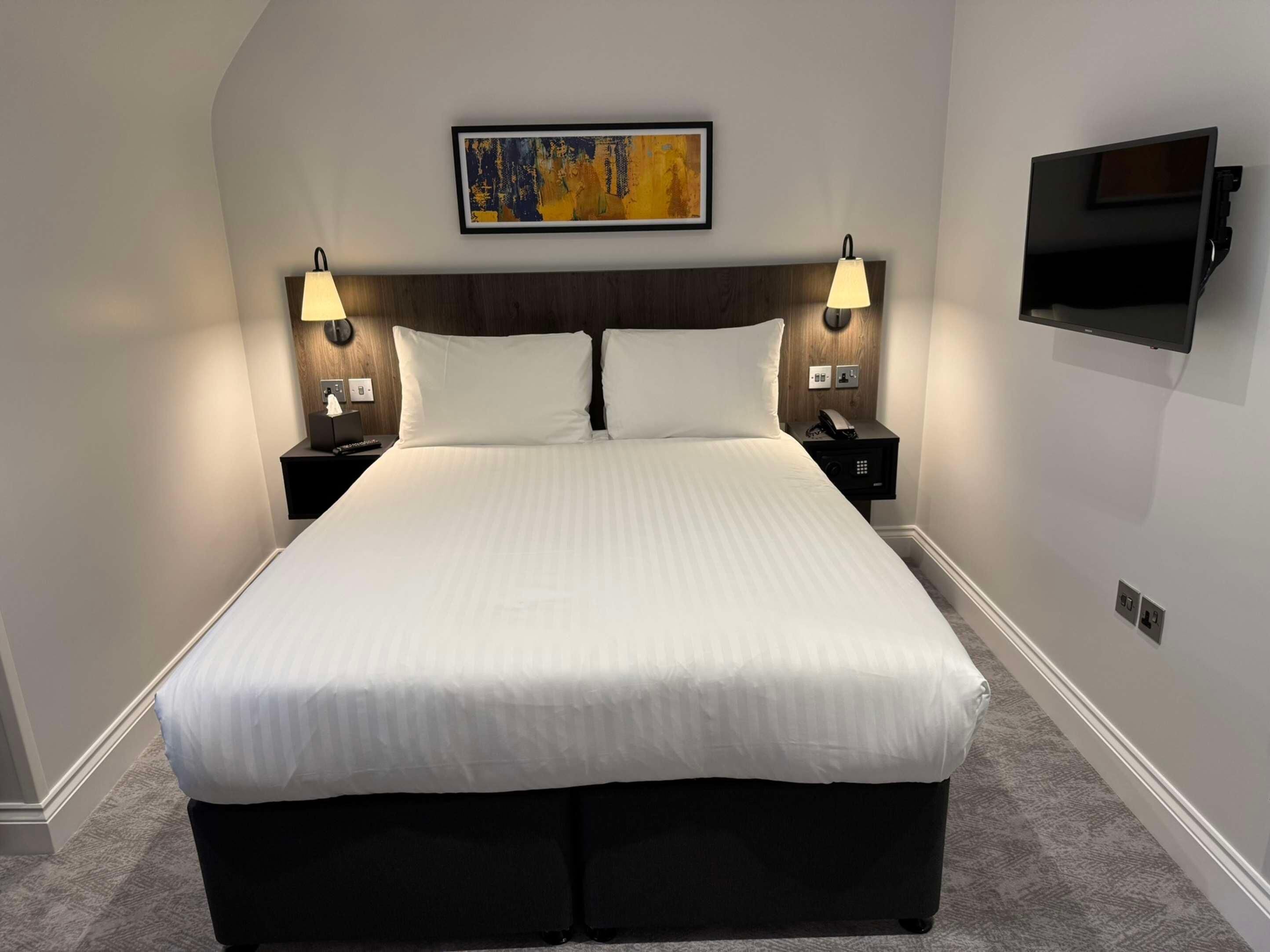 Una habitación de hotel con una cama grande, dos mesitas de noche con lámparas, un televisor montado y un cuadro en la pared.