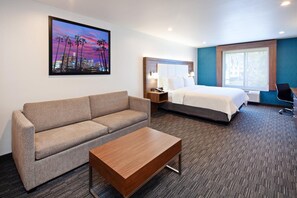 A hotel room with a bed, a sofa, a coffee table, a nightstand, and a window with a view of palm trees and a cityscape. - Best Western Kensington Olympia Hotel (London)