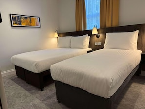 A hotel room with two beds, a nightstand, a lamp, a painting, and a window with curtains. - Best Western Kensington Olympia Hotel (London)