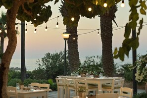 Un espace de restauration en plein air, avec des tables pour deux, entouré de palmiers et offrant une vue sur la mer.