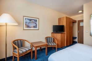 Ein Hotelzimmer mit Bett, Schreibtisch mit zwei Stühlen, Fernseher auf einem Holzschrank, einem gerahmten Bild an der Wand und einer Tür mit einem Schild.