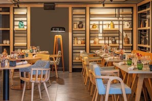 En restaurant med flere borde og stole, reoler med diverse genstande, og en midtergang.