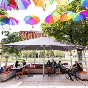 An outdoor seating area with tables and chairs, umbrellas, and a view of a building and trees. - Firstname Toulouse - JDV by Hyatt (Toulouse)