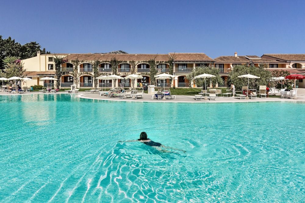 Una piscina con una persona che nuota, circondata da edifici di un hotel con balconi e ombrelloni.
