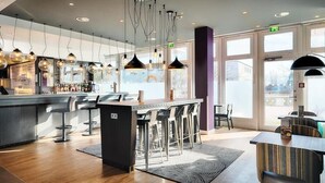 A modern bar area with a bar counter, stools, pendant lighting, and a view through large windows. - Azimut Hotel Munich (Munich)