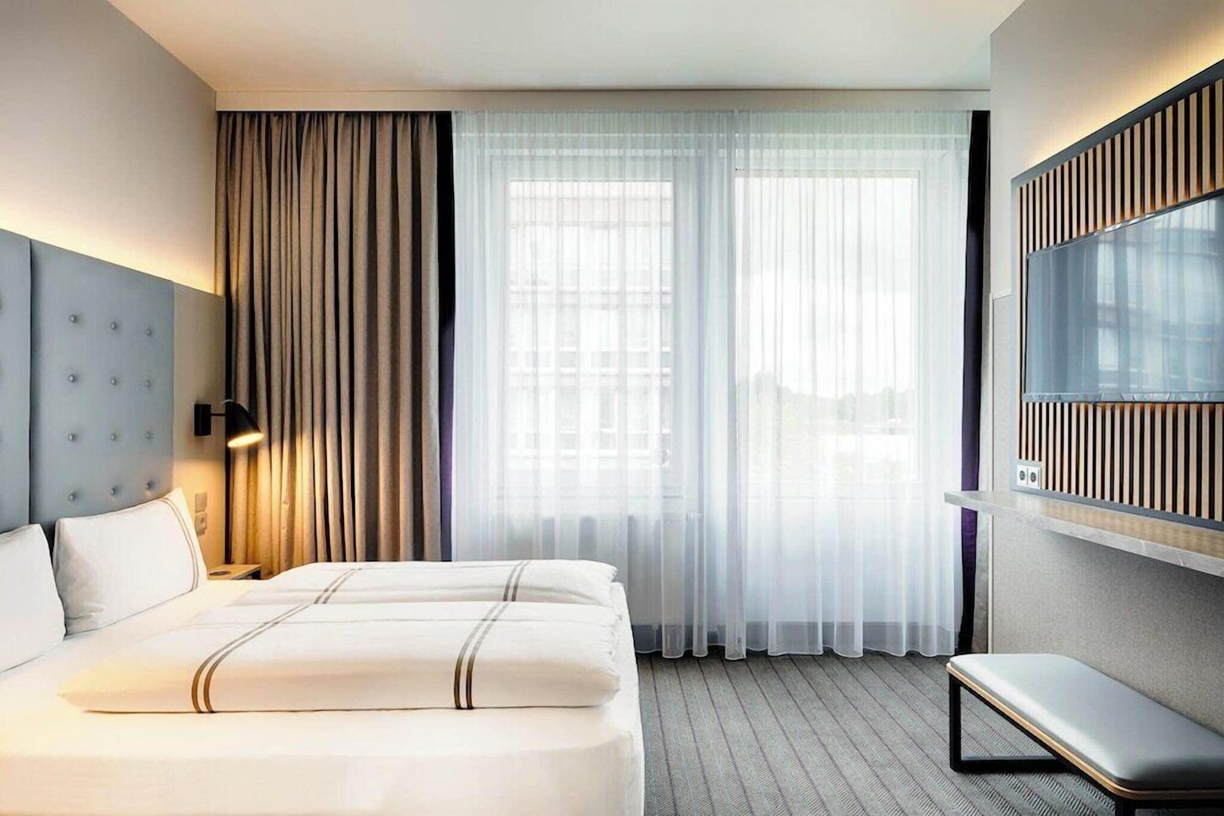 Ein Hotelzimmer mit einem großen Bett, einer Bank, einem Fernseher und einem Fenster mit Vorhängen.