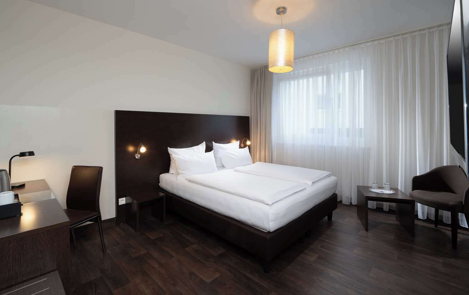 A hotel room with a large bed, a desk, a chair, a small table, and a lamp.