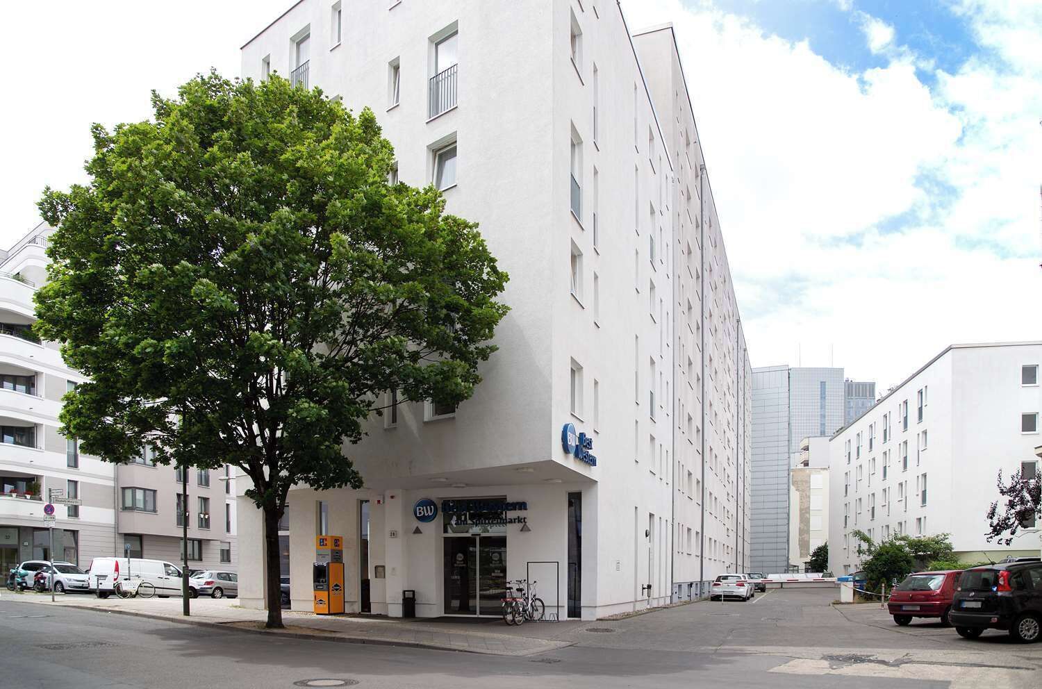 A modern building with a large tree in front, a sign for 'BW Hotel,' and several parked cars.