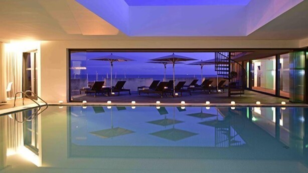 A modern indoor pool with lounge chairs and umbrellas, large windows offering a view of the ocean. - Pestana Cidadela Cascais (Cascais)