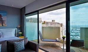 Una camera d'albergo moderna con un balcone, un letto, una poltrona e vista sul porto turistico.