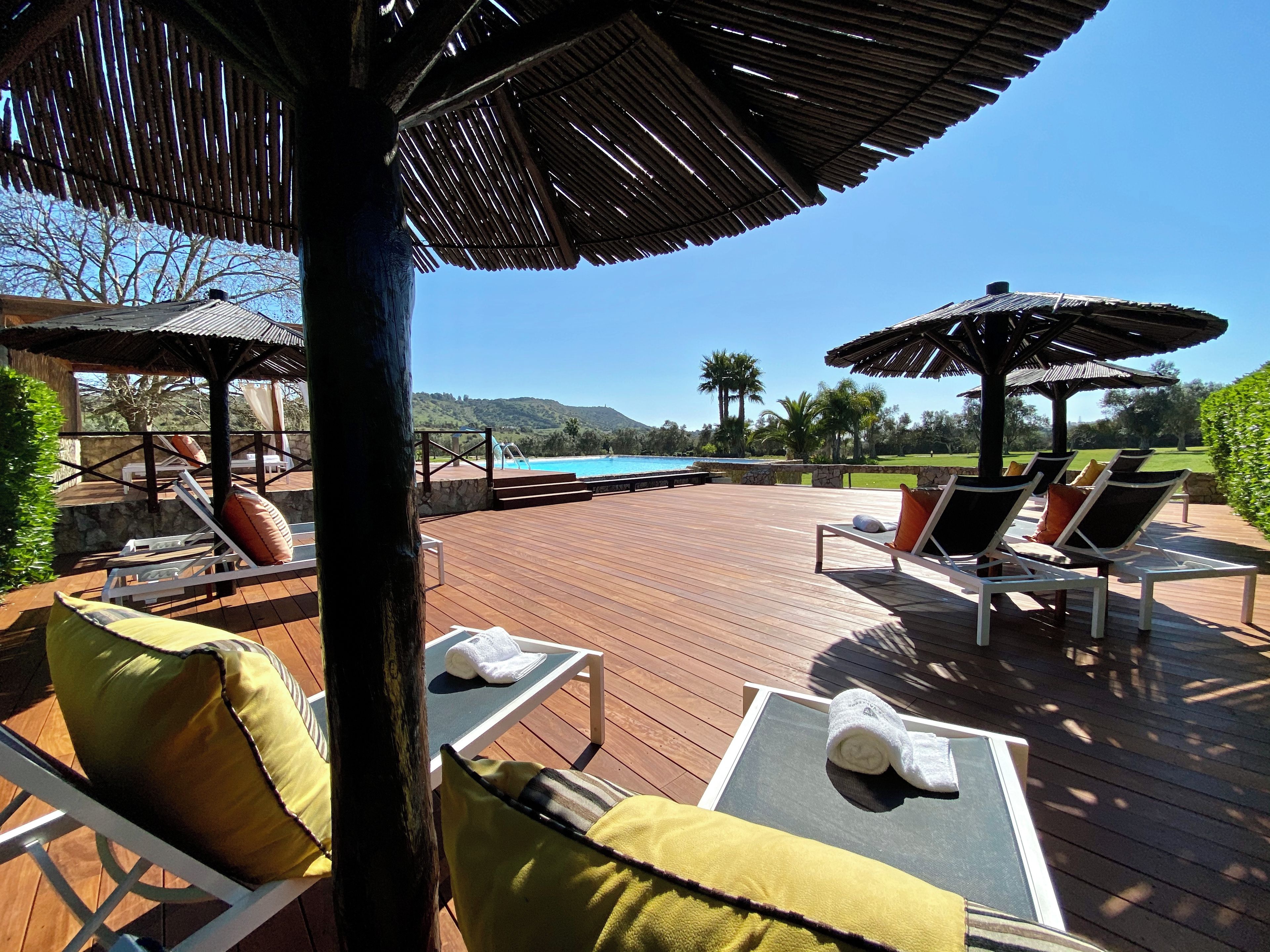 Une terrasse en bois, dotée de chaises longues et de parasols, qui offre une vue sur une piscine et un espace paysager aménagé.