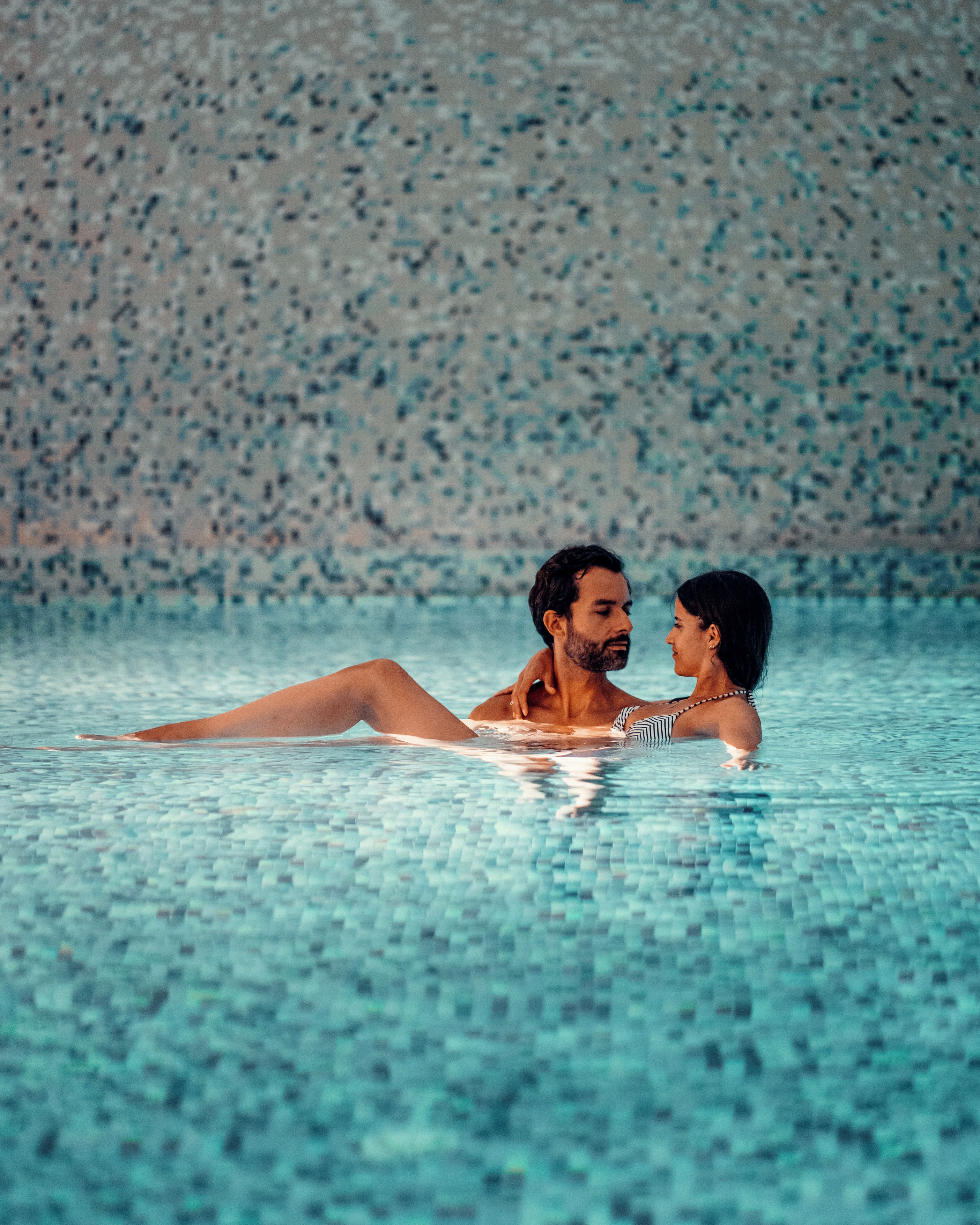 Una pareja recostada en una piscina azul cristalina con un fondo de azulejos con manchas.