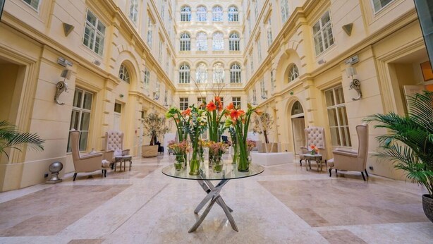 Ein geräumiger Innenhof mit Glastisch, umgeben von Stühlen, einer zentralen Blumenkomposition und einer hohen Decke mit Bogenfenstern.