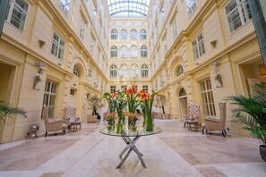 A spacious atrium with a glass-top table surrounded by chairs, a central floral arrangement, and a high ceiling with arched windows. - Grand Palace Brno Hotel (Brno)