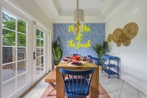 A dining area with a table set for four, a blue chair, a blue side table, and a wall with a tropical pattern and a neon sign. - Sleeps 10 • Heated Pool • Game Room • Hot Tub (Largo)