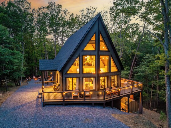 A cabin with a large deck, multiple windows, and a gravel driveway surrounded by trees.