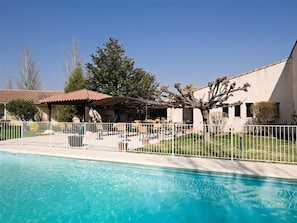 A swimming pool with a covered patio area, a fenced yard, and a building with a sloped roof.