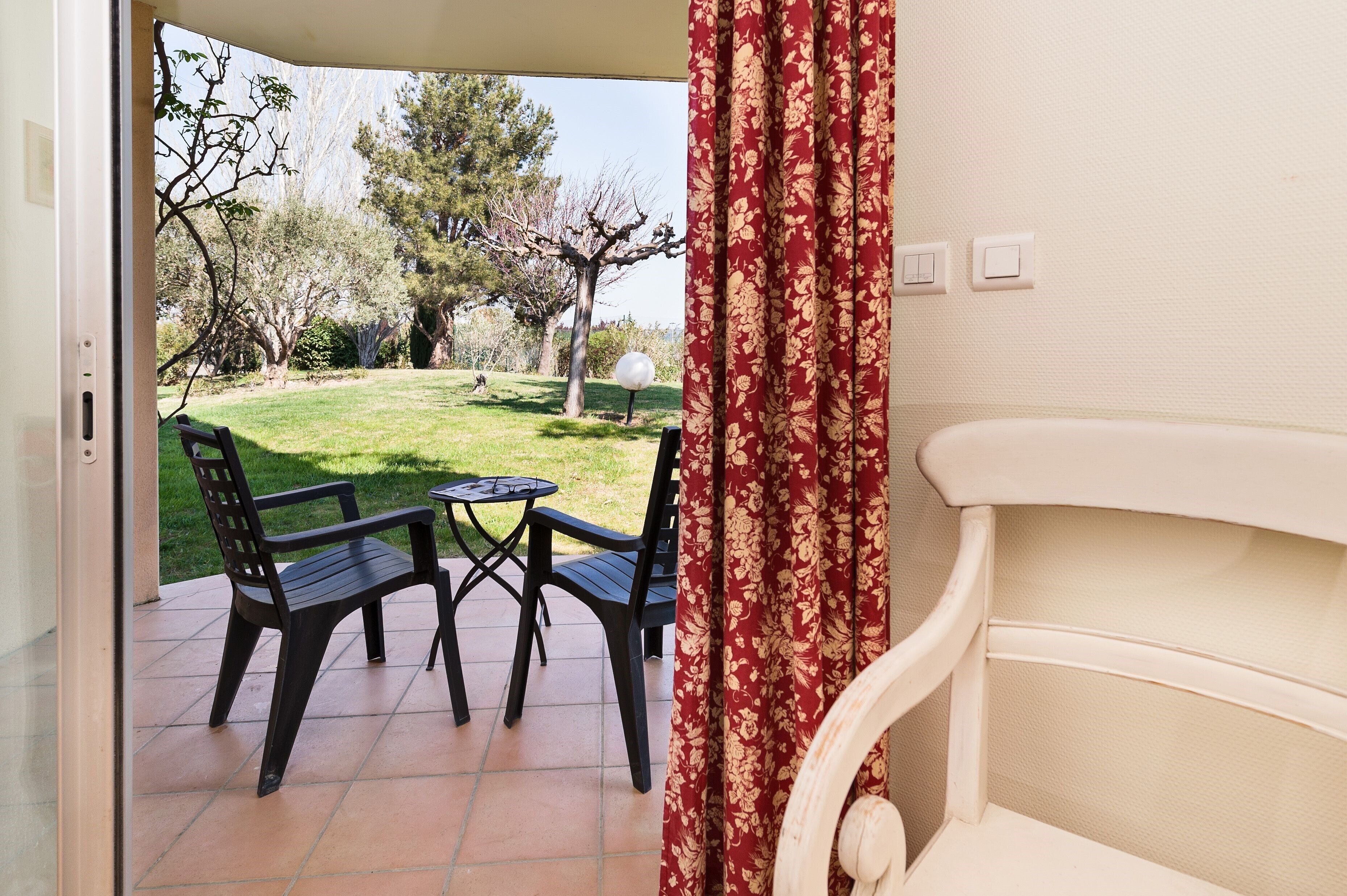 Une terrasse ouverte avec une table et des chaises, une vue sur un jardin avec des arbres et une pelouse, et un mur blanc avec un rideau.