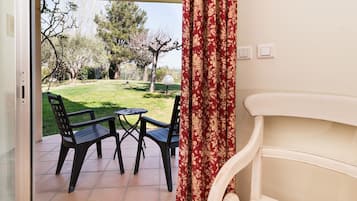 Une terrasse ouverte avec une table et des chaises, une vue sur un jardin avec des arbres et une pelouse, et un mur blanc avec un rideau.