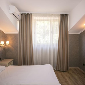 A hotel room with a bed, a window with sheer curtains, a wall-mounted air conditioner, and two wall sconces. - Hotel Royal Bay (Nessebar)