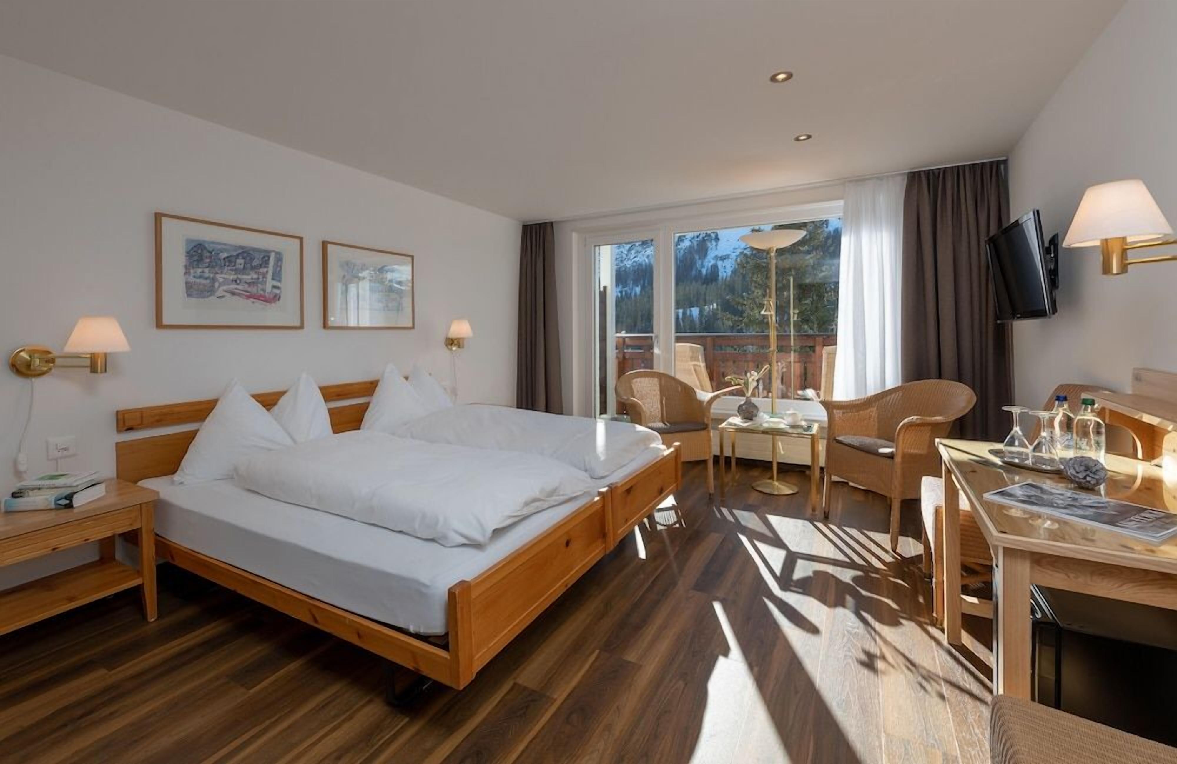Une chambre d’hôtel comprenant un lit, des tables de chevet, un bureau avec des chaises, une télévision et des tableaux encadrés accrochés au mur.
