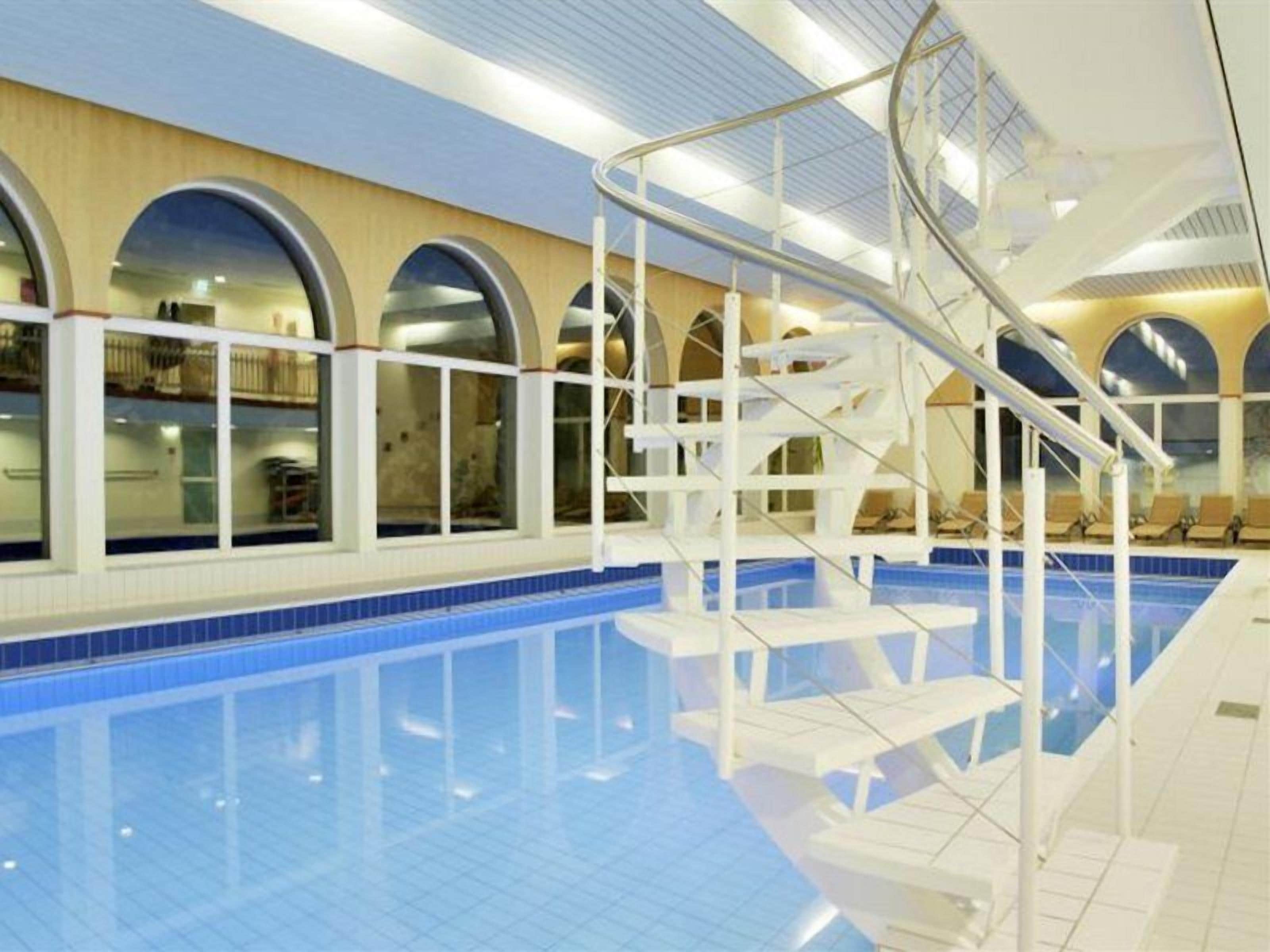 Une piscine intérieure dotée d’un plongeoir, de fenêtres en arc de cercle et d’un sol carrelé.