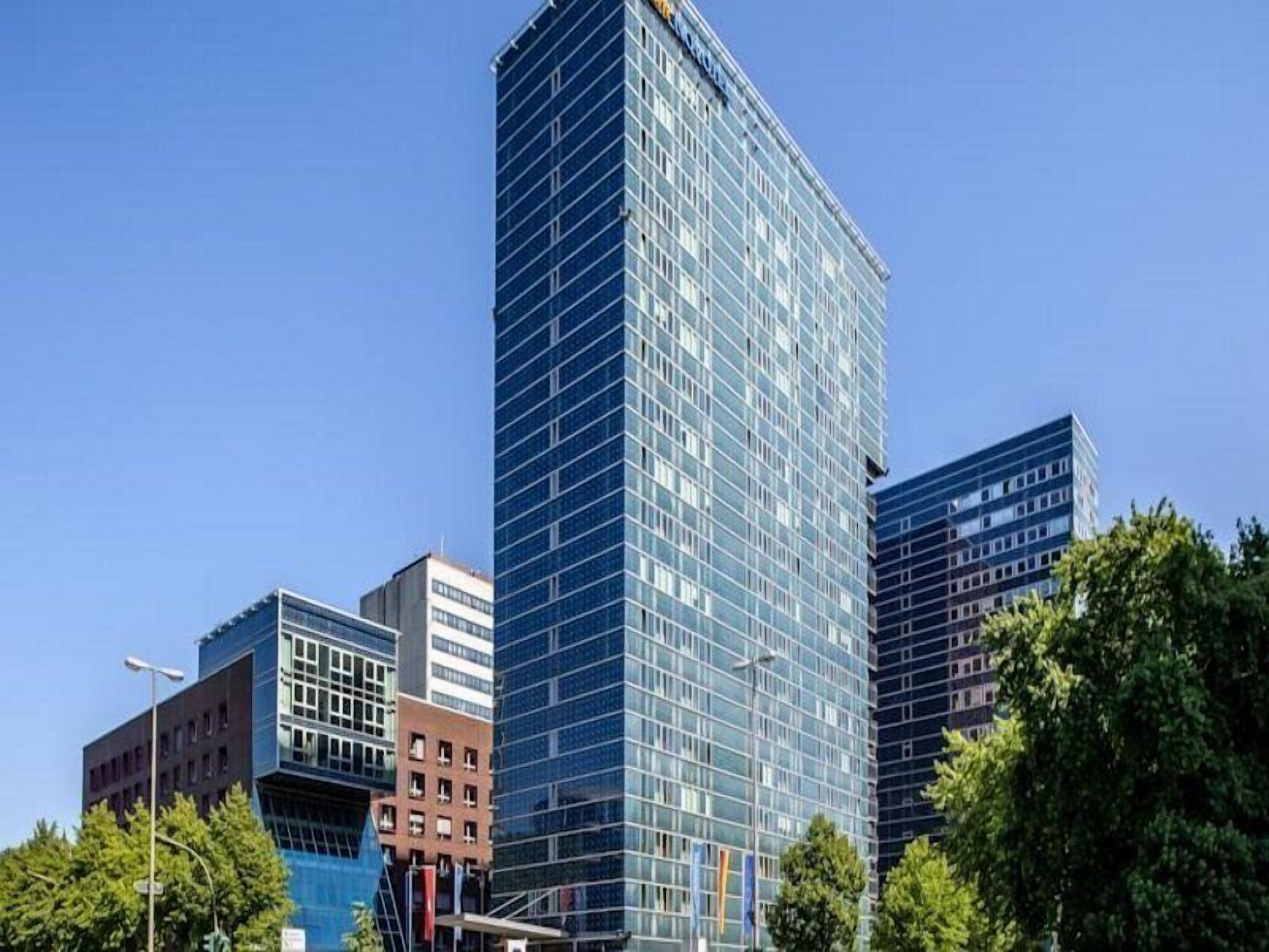 Un moderno edificio de gran altura con fachada de vidrio, rodeado de otros edificios y árboles.