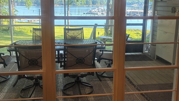 Uitzicht door een glazen deur, met een bordje 'Welcome to the Lake', een tafel met stoelen, en een meer met aangemeerde boten.
