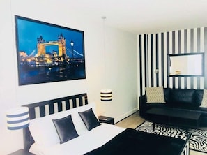 Una habitación de hotel con una cama, pared a rayas blancas y negras, un sofá negro, un tapete estampado de cebra, un cuadro de un puente y una lámpara.