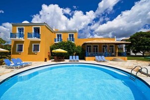 A pool area with a clear blue pool, a yellow building with balconies, and a covered seating area.