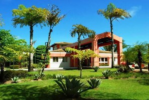 Um edifício moderno com telhado plano, rodeado por vegetação bem cuidada e palmeiras.