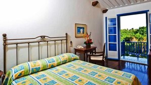 A bedroom with a bed, a bedside table, a chair, a framed picture on the wall, and a balcony with a view of greenery. - Sauipe Pousadas (Mata de São João)