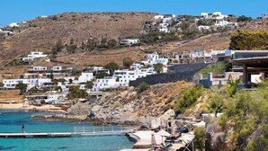 A coastal hotel with beachside lounge chairs and umbrellas, a rocky shoreline, and white buildings on a hillside. - Saint John Hotel Villas and Spa (Mykonos)