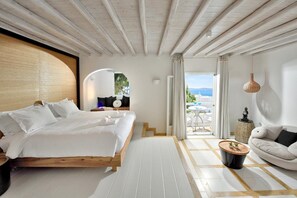 A modern hotel room with a wooden bed, a round coffee table, a sofa, and a view of the outdoors. - Saint John Hotel Villas and Spa (Mykonos)