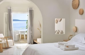 A hotel room with a bed, a table with chairs, a view of the sea, and a bathroom with a mirror and towel rack. - Saint John Hotel Villas and Spa (Mykonos)