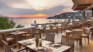 An outdoor dining area with tables and chairs, overlooking a body of water and a hilly landscape during sunset. - Saint John Hotel Villas and Spa (Mykonos)