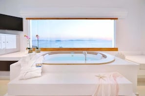 A modern bathroom with a large bathtub, a vanity with a mirror, and a window offering a view of the sea. - Saint John Hotel Villas and Spa (Mykonos)