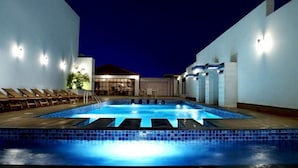 A swimming pool with lounge chairs, a sun terrace, and a building with a glass facade. - Savoy Grande Hotel & Spa (Manama)