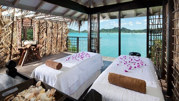 Una habitación con dos camas, una terraza de madera, vista al mar y un muro de paja.