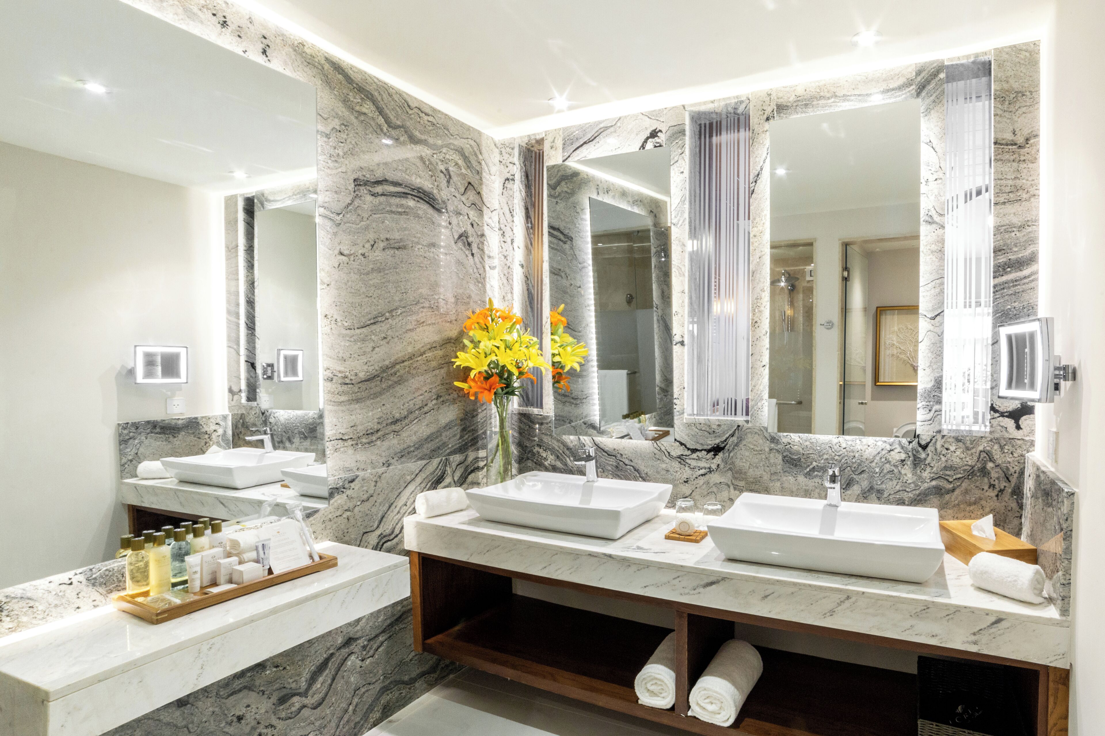 A modern bathroom with two sinks, marble walls, a large mirror, and a vase with flowers.
