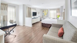 Ein Hotelzimmer mit Bett, Sofa, Schreibtisch mit Computer, Fernseher und großem Fenster mit Vorhängen.
