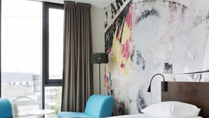 Ein Hotelzimmer mit einem Bett, einem blauen Sessel, einem kleinen Tisch und Blick auf Gebäude durch ein Fenster.